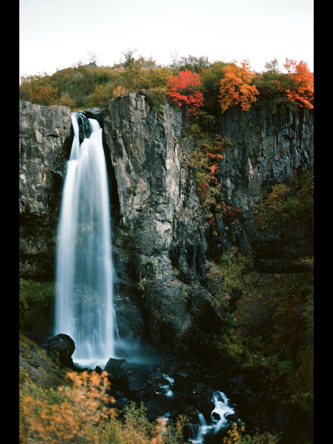 A tall waterfall cascades down a rocky cliff with autumn foliage at the top.