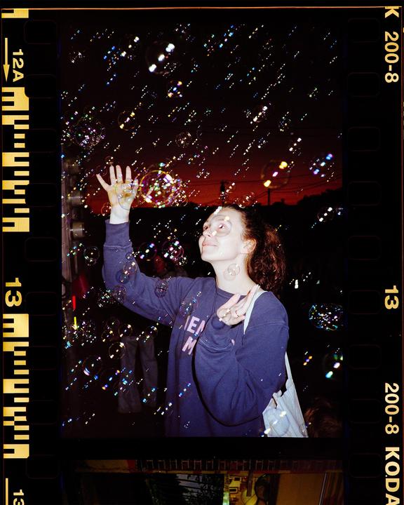 A woman with curly hair looks up at numerous bubbles floating around her against a dark sky with a red glow.