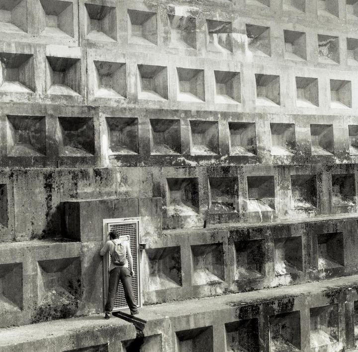 A man with a backpack stands in front of a metal door set into a concrete wall with many square indentations.