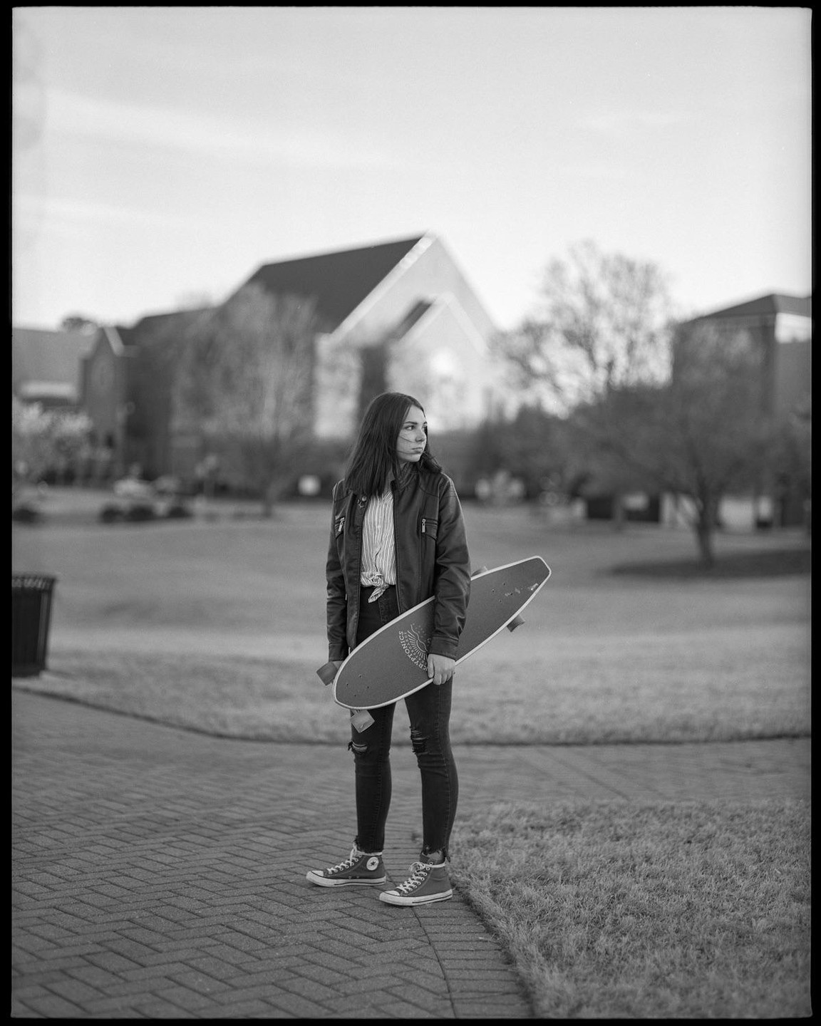 A young woman stands on a paved path holding a skateboard, looking to her right.