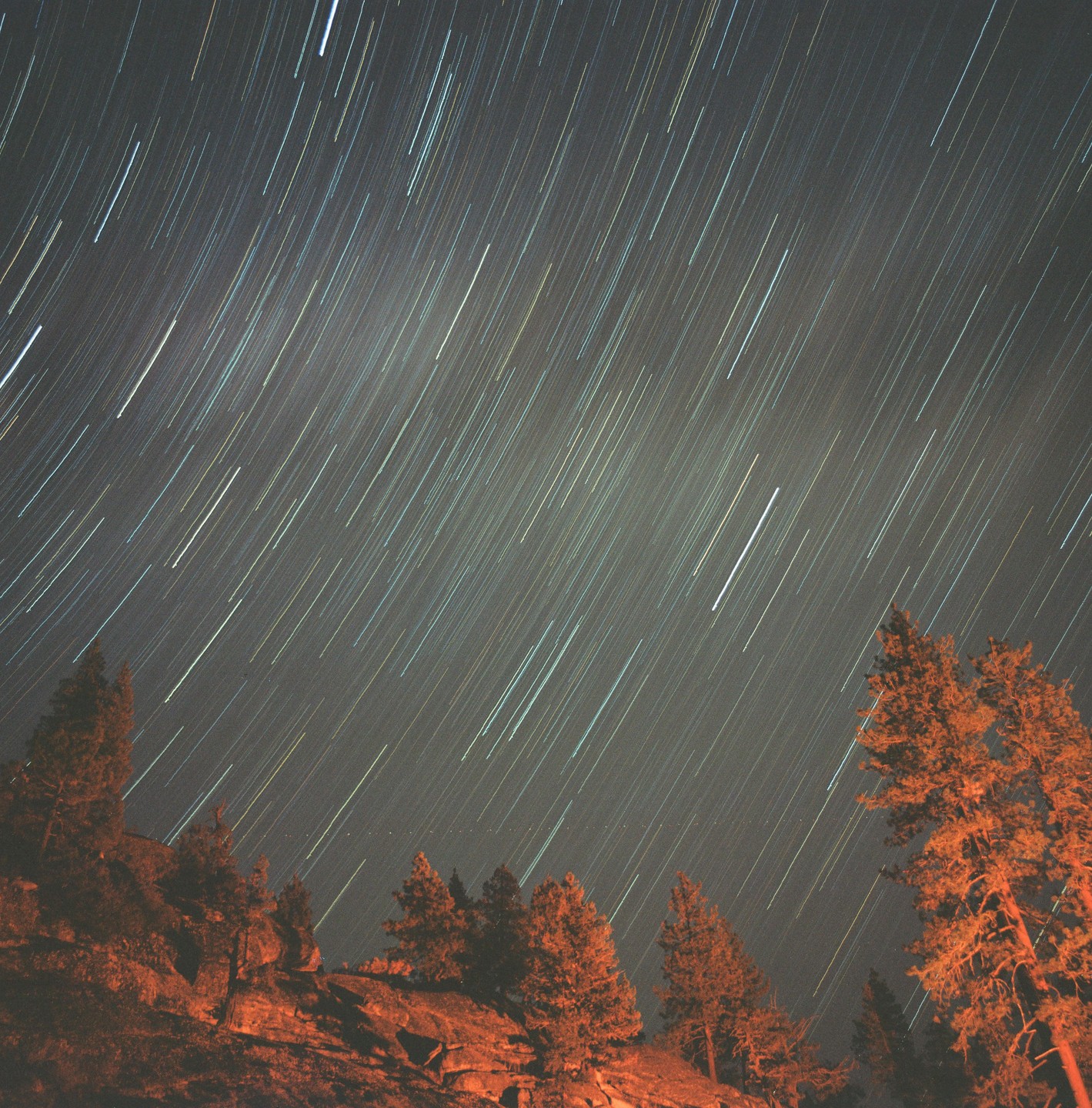 A long exposure photograph captures star trails above a rocky landscape with trees illuminated by an orange light.