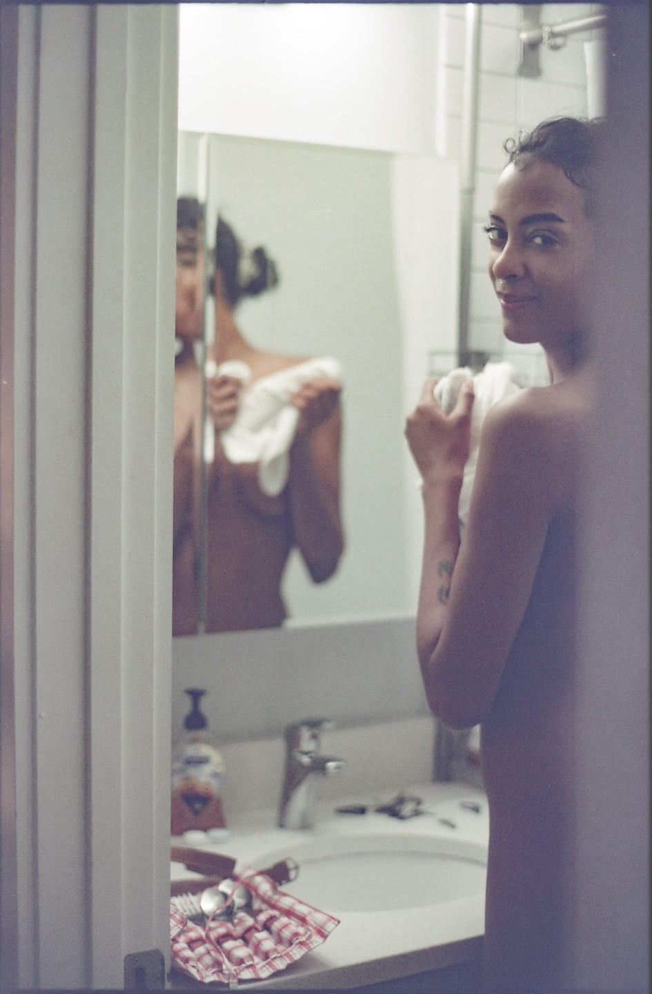A woman stands in a bathroom, drying herself with a towel, with her reflection visible in the mirror.