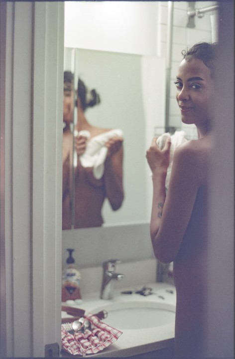 A woman stands in a bathroom, drying herself with a towel, with her reflection visible in the mirror.