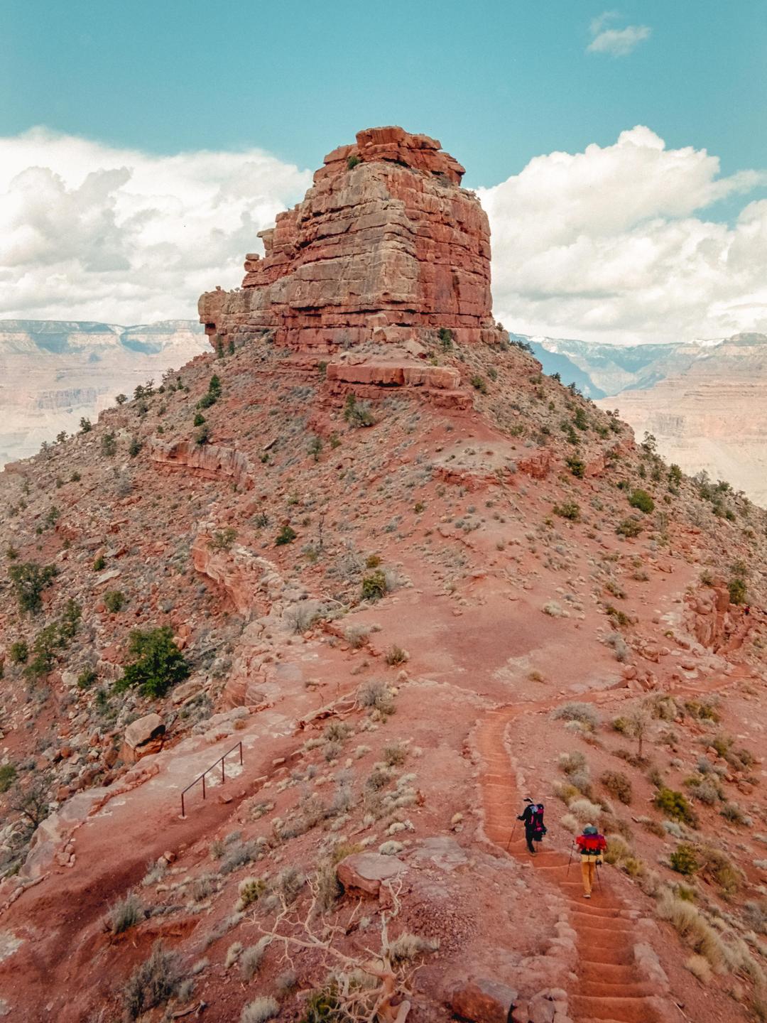 Two people hike on a winding trail leading up to a large red rock formation under a partly cloudy sky.