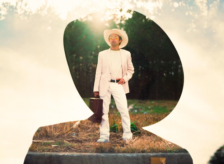 A man in a white suit, cowboy hat, and sunglasses stands holding a briefcase in a double exposure.