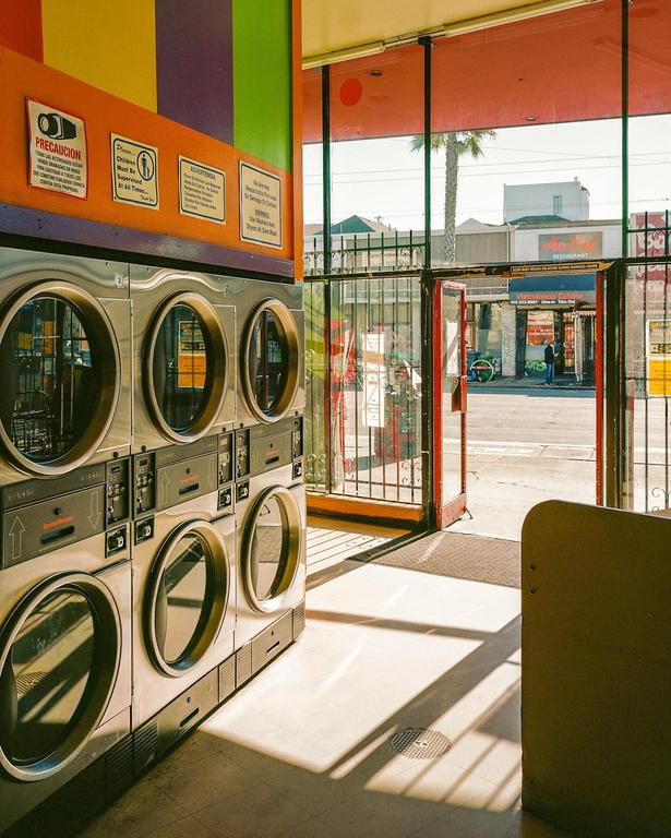 Sunlight streams into a laundromat with a row of washing machines and an open door leading to a street.