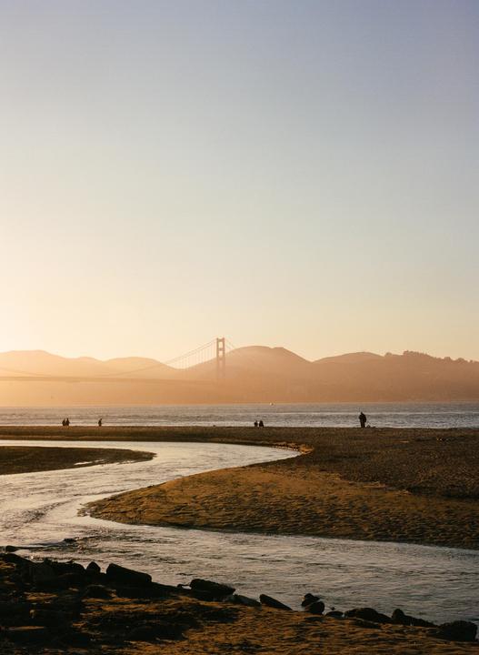 A winding river flows into a body of water with a bridge and mountains in the distance during golden hour.