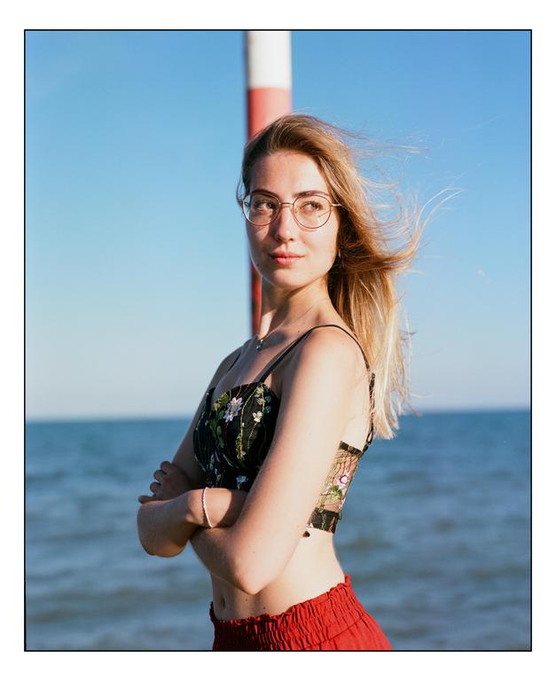 A woman with glasses and long hair stands with her arms crossed in front of the sea.