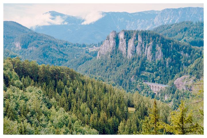 A blue train crosses a stone viaduct in a valley surrounded by mountains and dense forest.