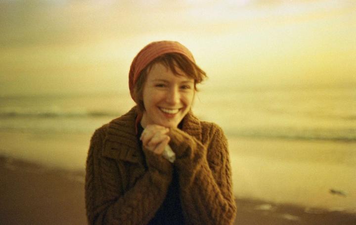 A woman in a brown sweater and red headscarf smiles at the camera on a beach.