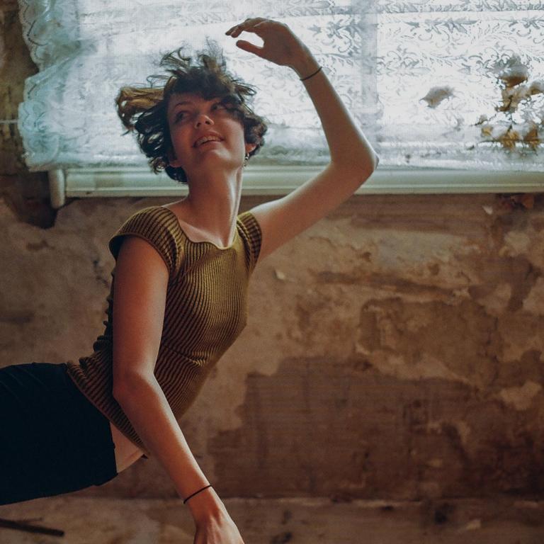 A woman with short hair and a striped shirt is posing with her arms raised in front of a window with a lace curtain.