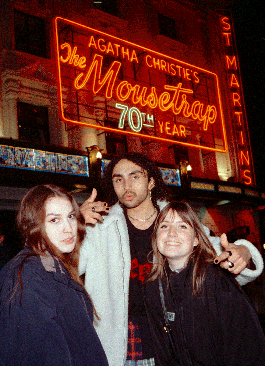 A man and two women pose in front of a neon sign for "Agatha Christie's The Mousetrap" at night.