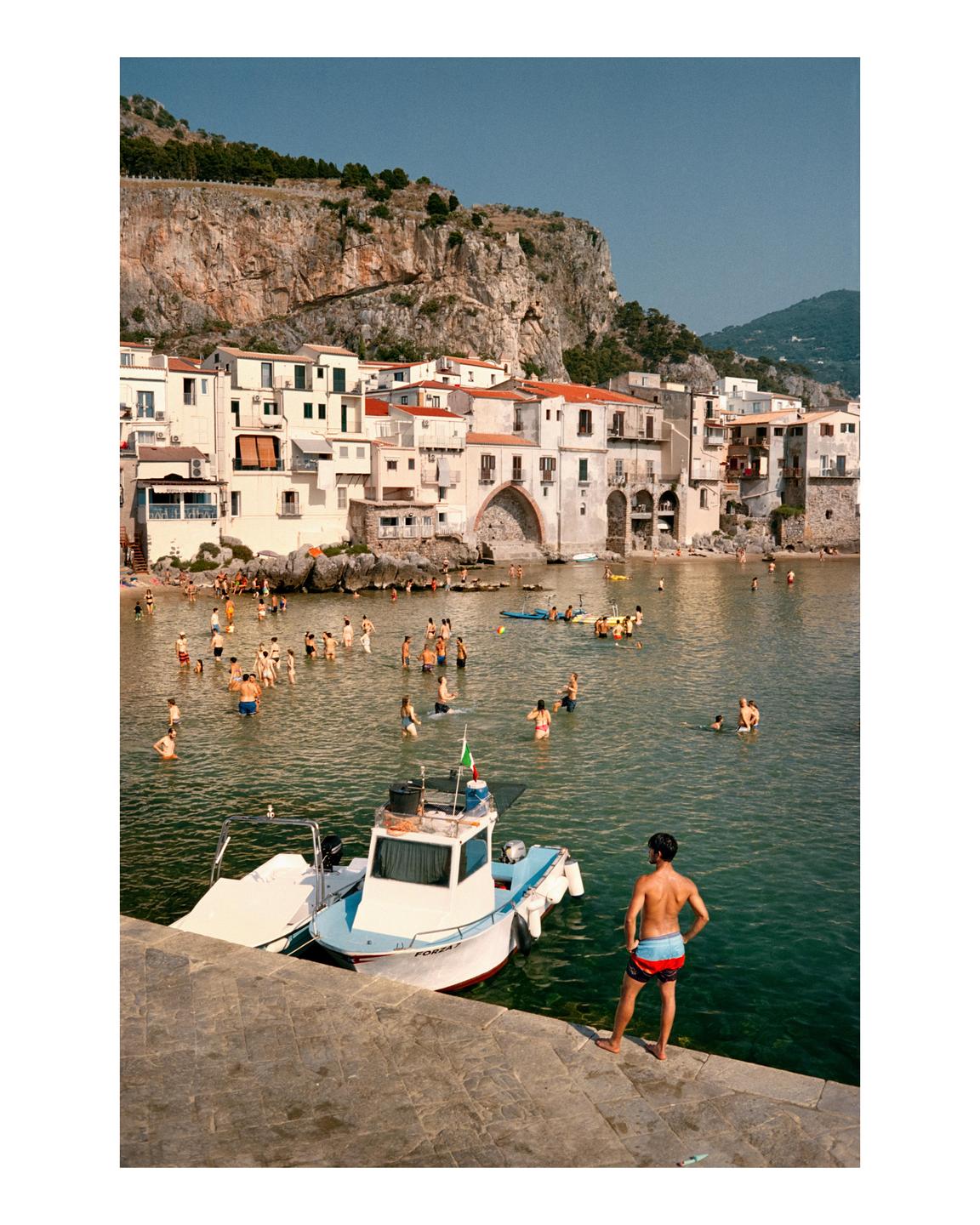 A man stands on a stone pier looking out at a bay filled with swimmers and boats, with a town built into a cliffside in the background.