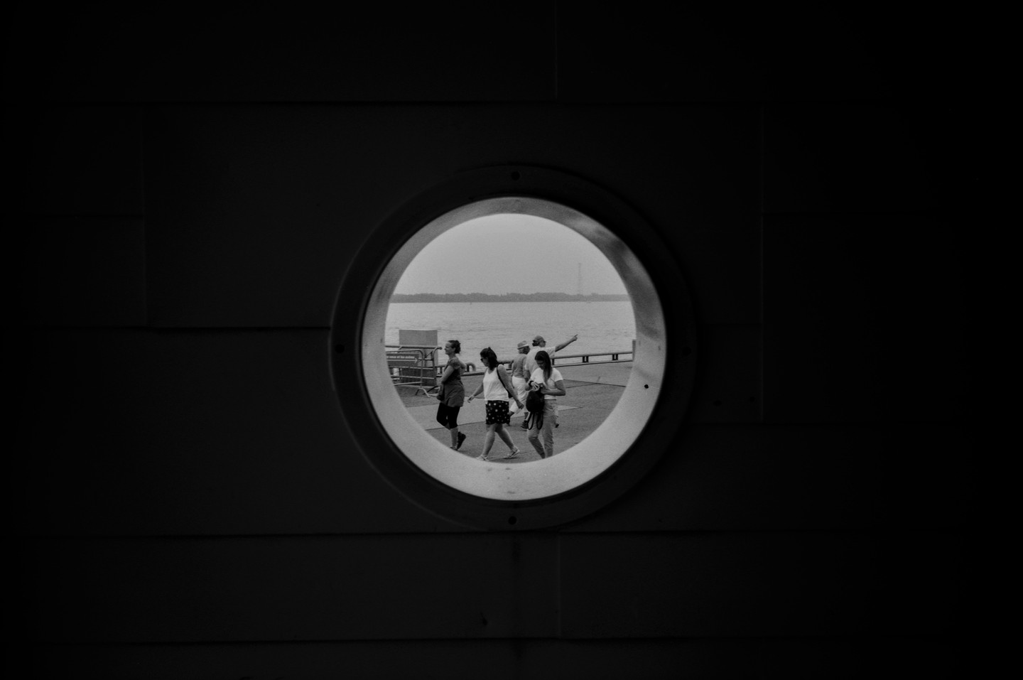 A group of people walk on a dock seen through a porthole, with water and a distant shore visible in the background.