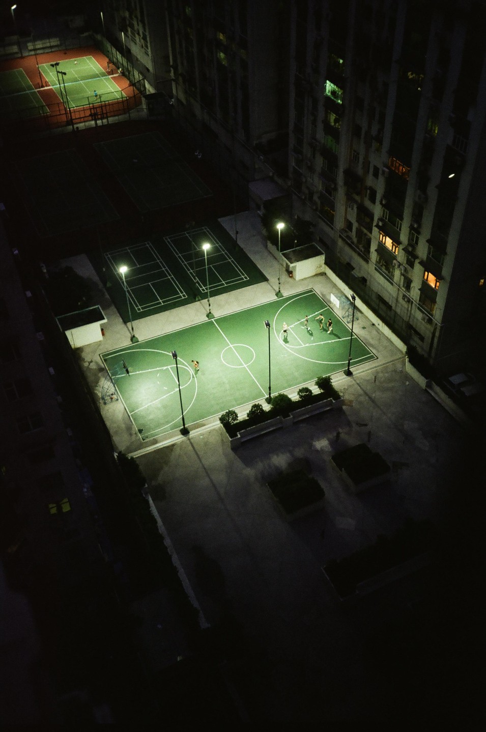 An aerial view shows several people playing basketball on a brightly lit court at night, surrounded by other courts and a tall building.