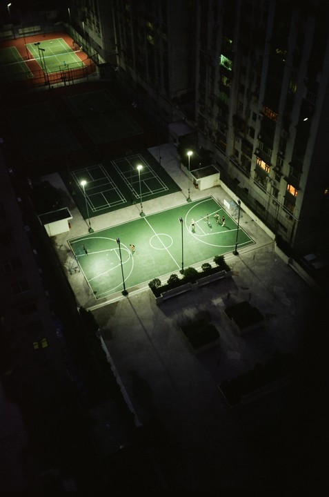 An aerial view shows several people playing basketball on a brightly lit court at night, surrounded by other courts and a tall building.