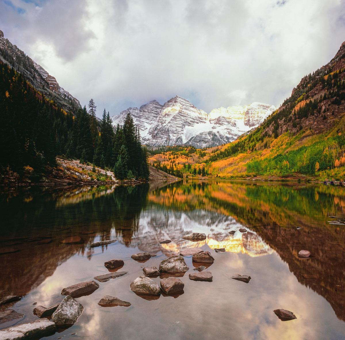 Snow-capped mountains and autumn trees reflect in a lake with rocks in the foreground.
