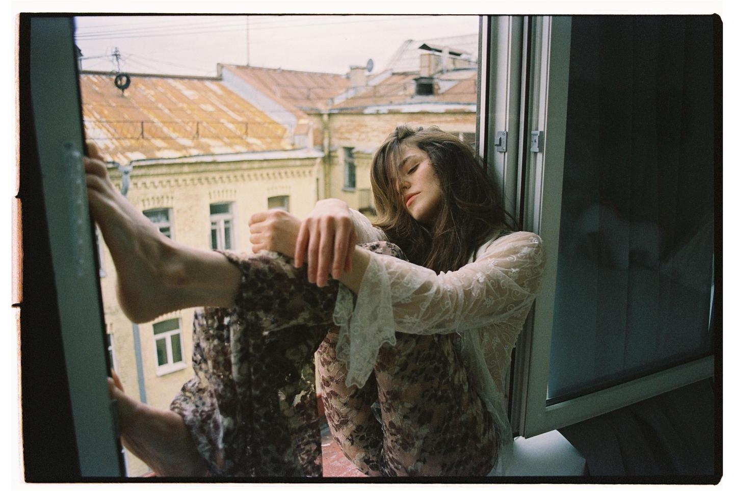 A woman with long hair and a lace top sits in an open window overlooking a city.