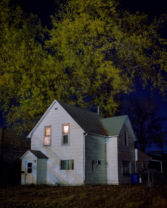 A white house with illuminated windows stands under a large tree with yellow foliage at night.