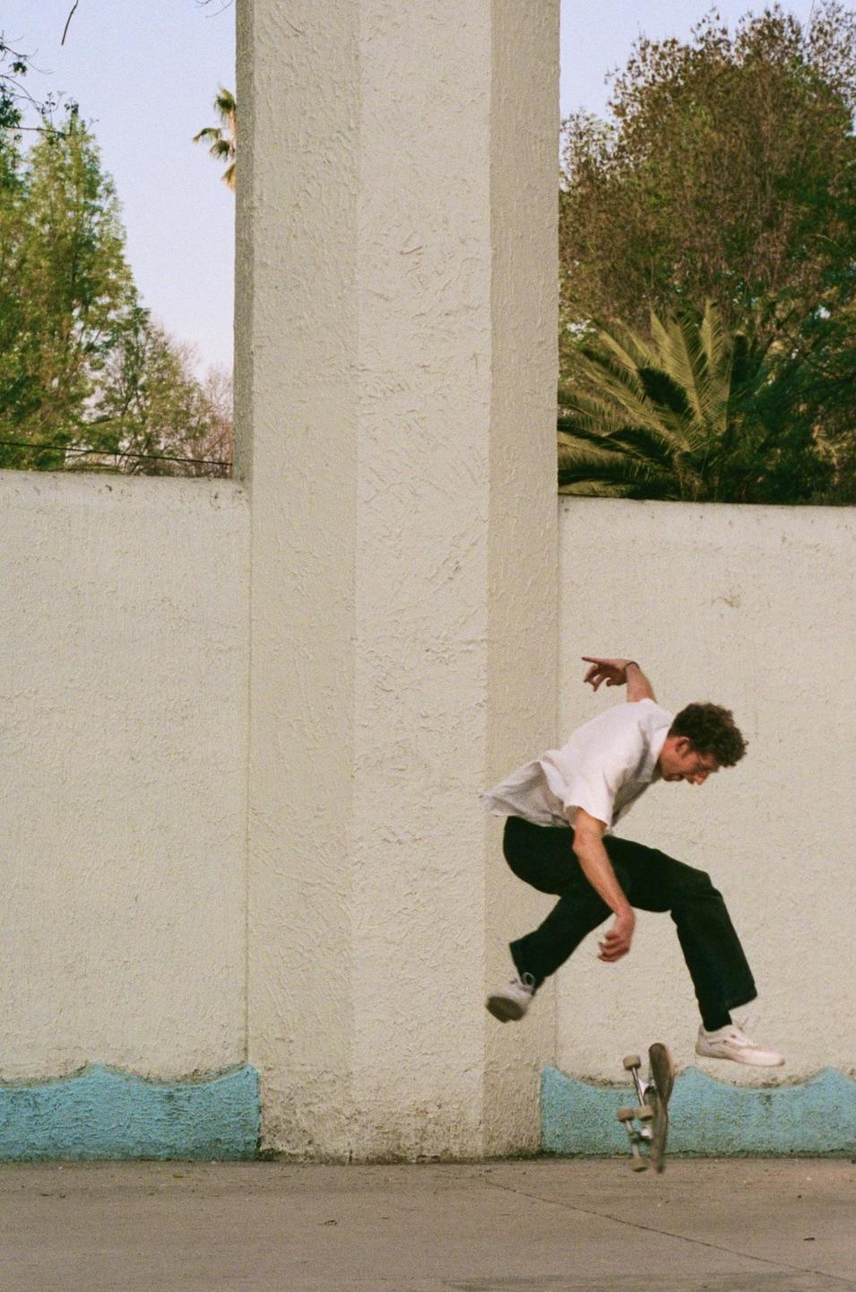 A man in a white shirt and black pants is captured mid-air while skateboarding next to a textured wall with a blue base.