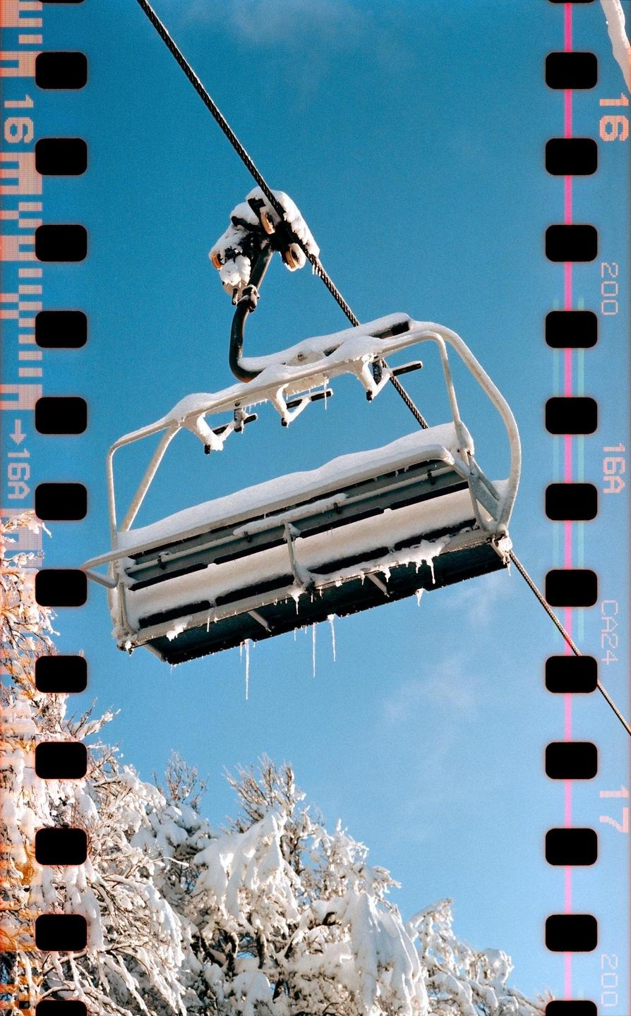 A snow-covered ski lift chair hangs above snow-laden trees against a clear blue sky.