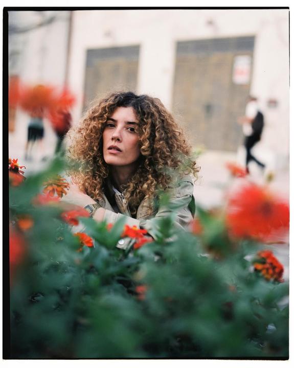 A woman with curly hair looks directly at the viewer from behind a bush with red flowers.