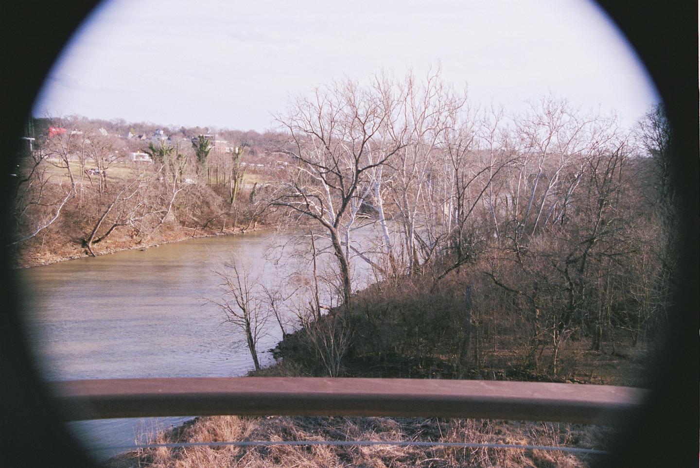 A river flows past a wooded bank with bare trees and a distant town under a bright sky, viewed through a circular frame.