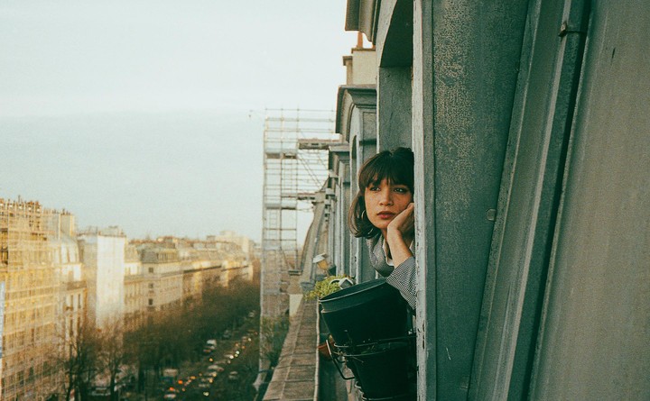 A woman with bangs looks out from a balcony over a city street.