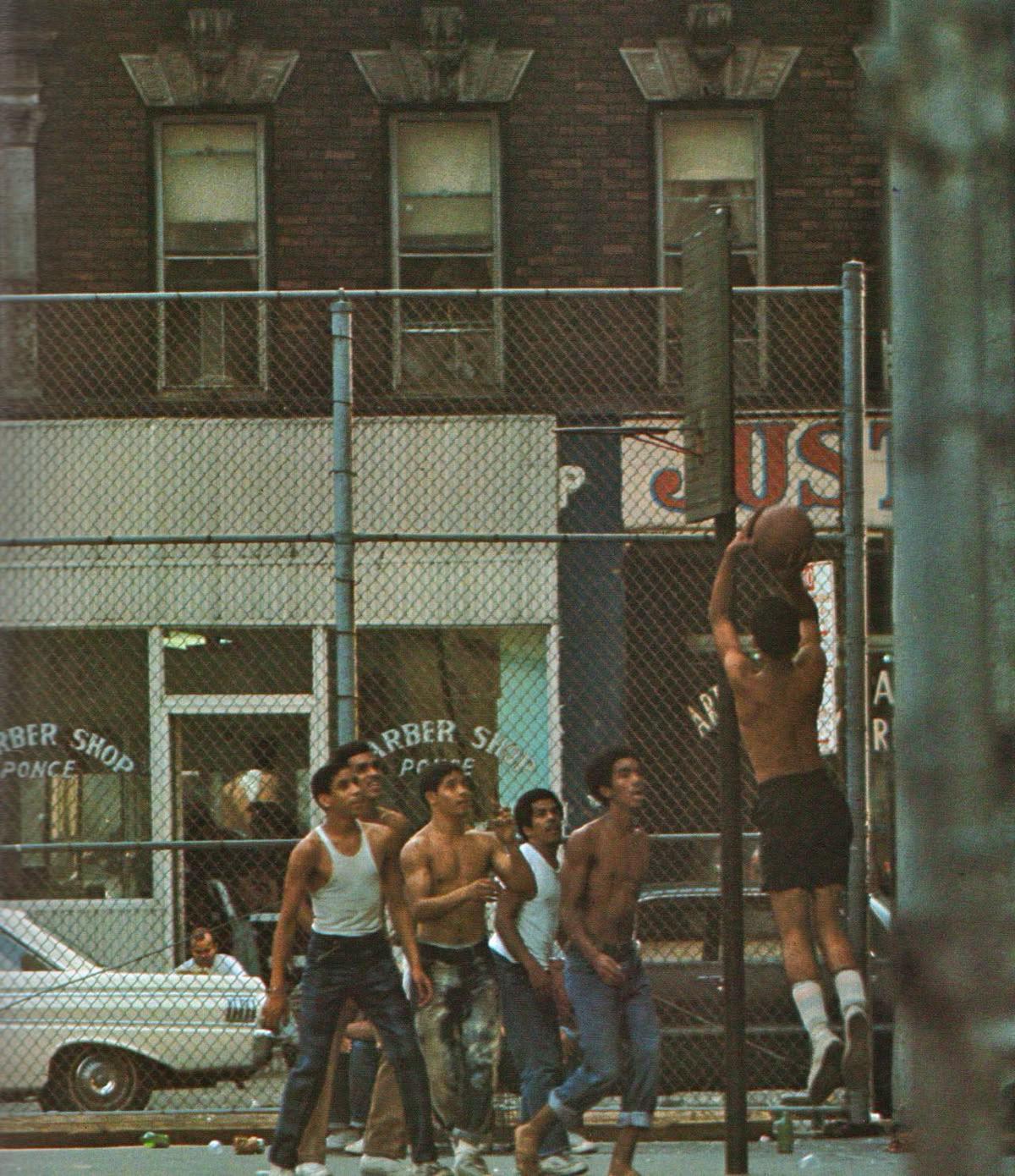 A group of young men play basketball on a street court in front of a barber shop.