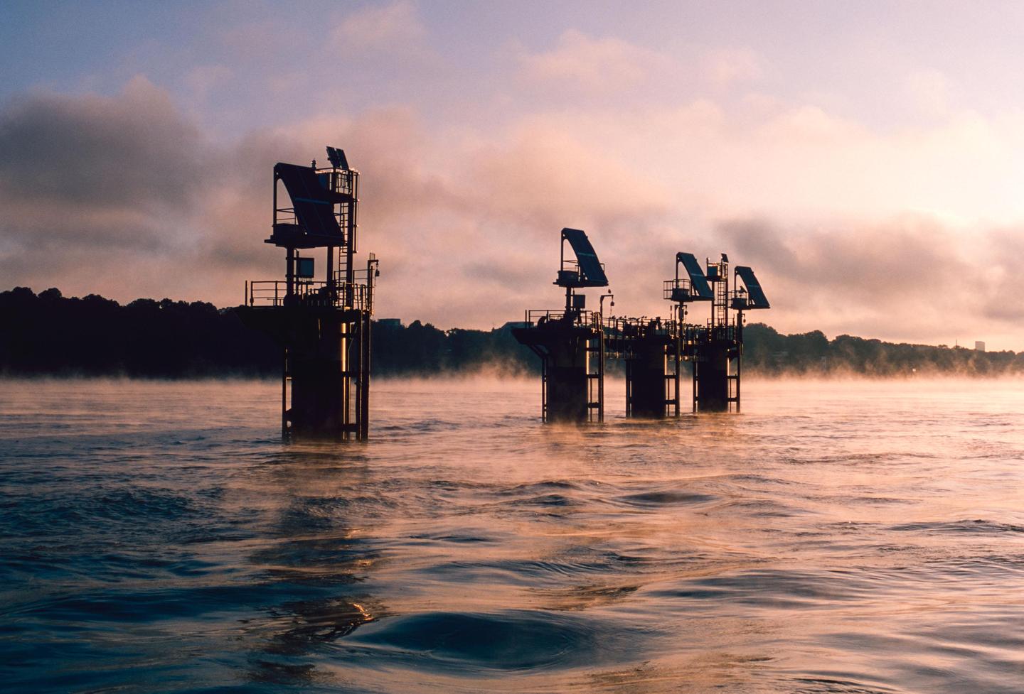 Several dark industrial structures stand in a misty body of water under a cloudy sky at dusk.