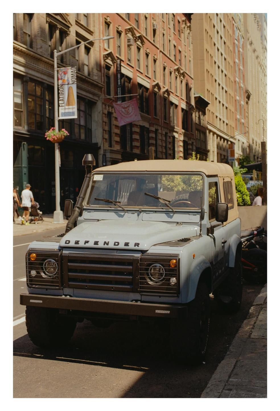 A light blue Land Rover Defender is parked on a city street with tall buildings in the background.