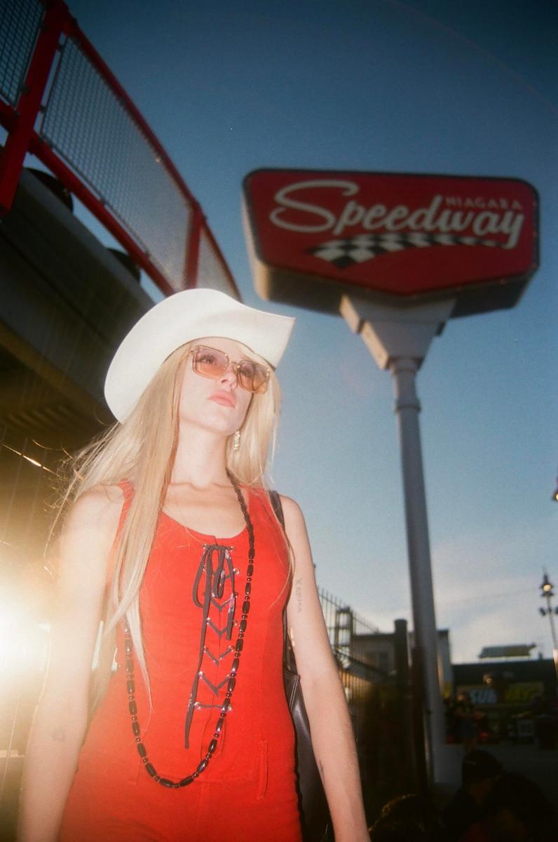 A woman wearing a cowboy hat and sunglasses stands in front of a Speedway sign.