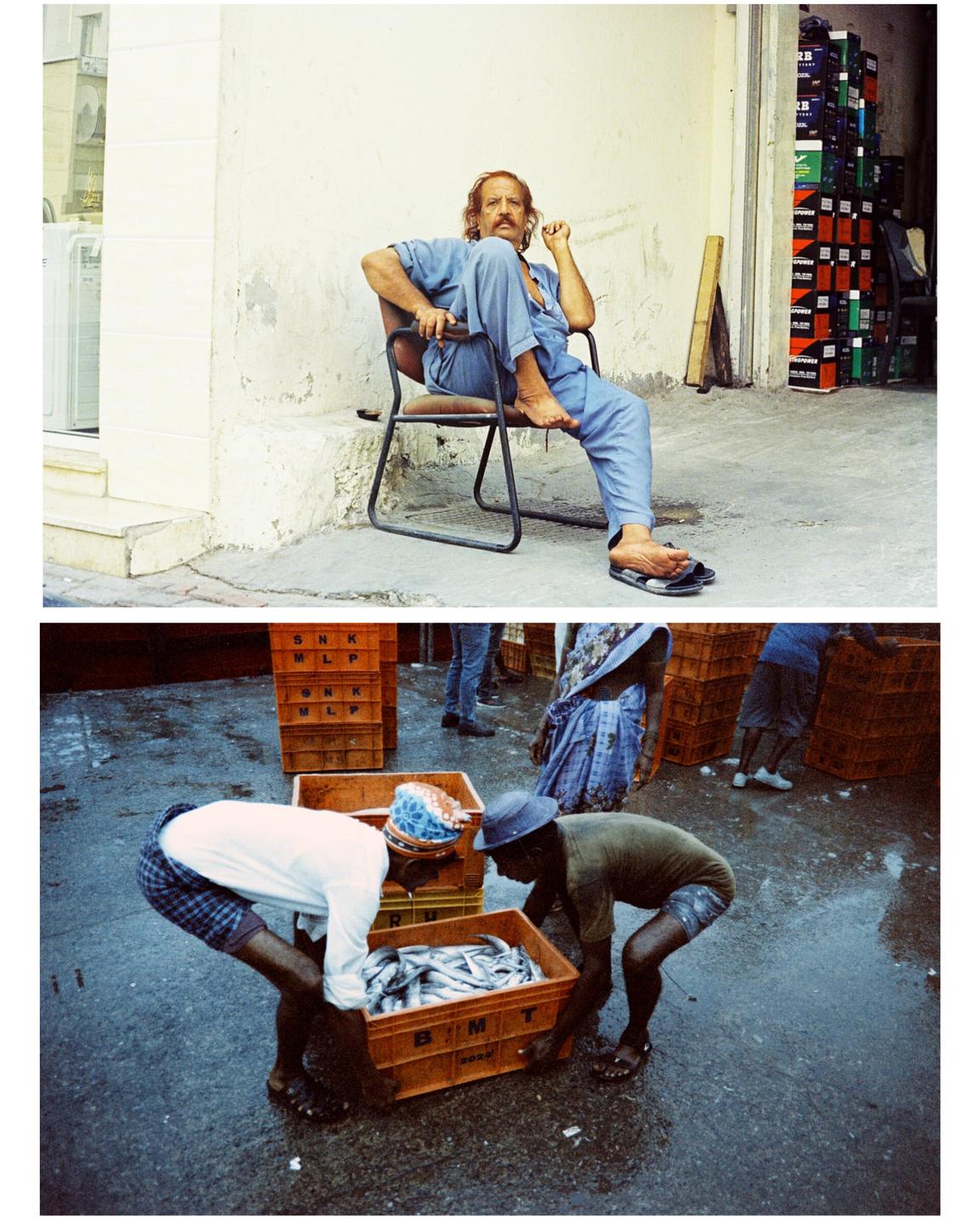A man with a mustache sits on a chair next to a building, and two men carry a crate of fish on a wet street.
