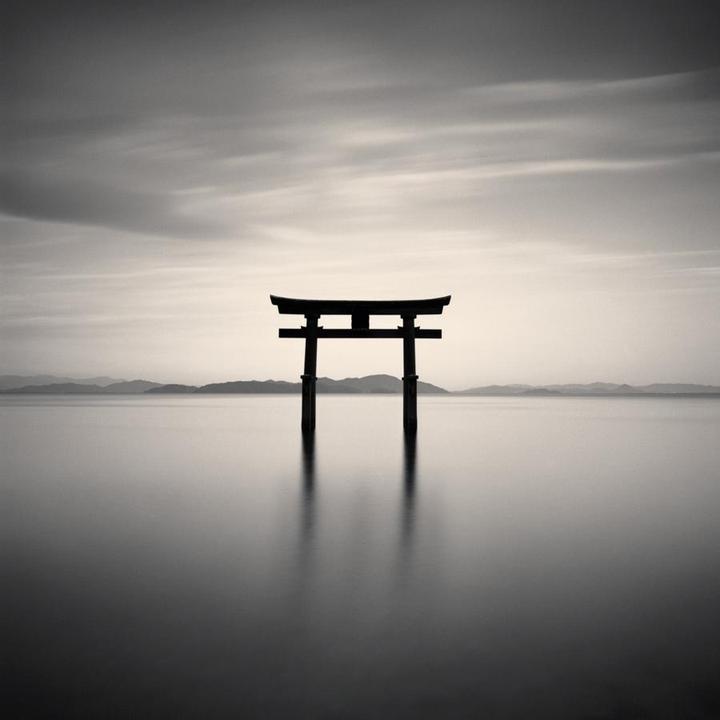 A torii gate stands in the middle of a calm lake with mountains in the distance under a cloudy sky.