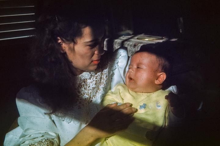 A woman in a white lace top holds a sleeping baby in a yellow outfit.