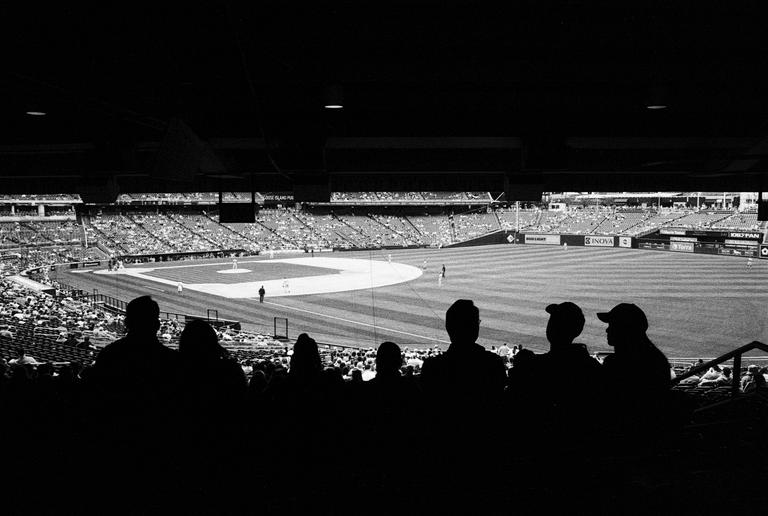 Silhouetted spectators watch a baseball game from the stands of a stadium.