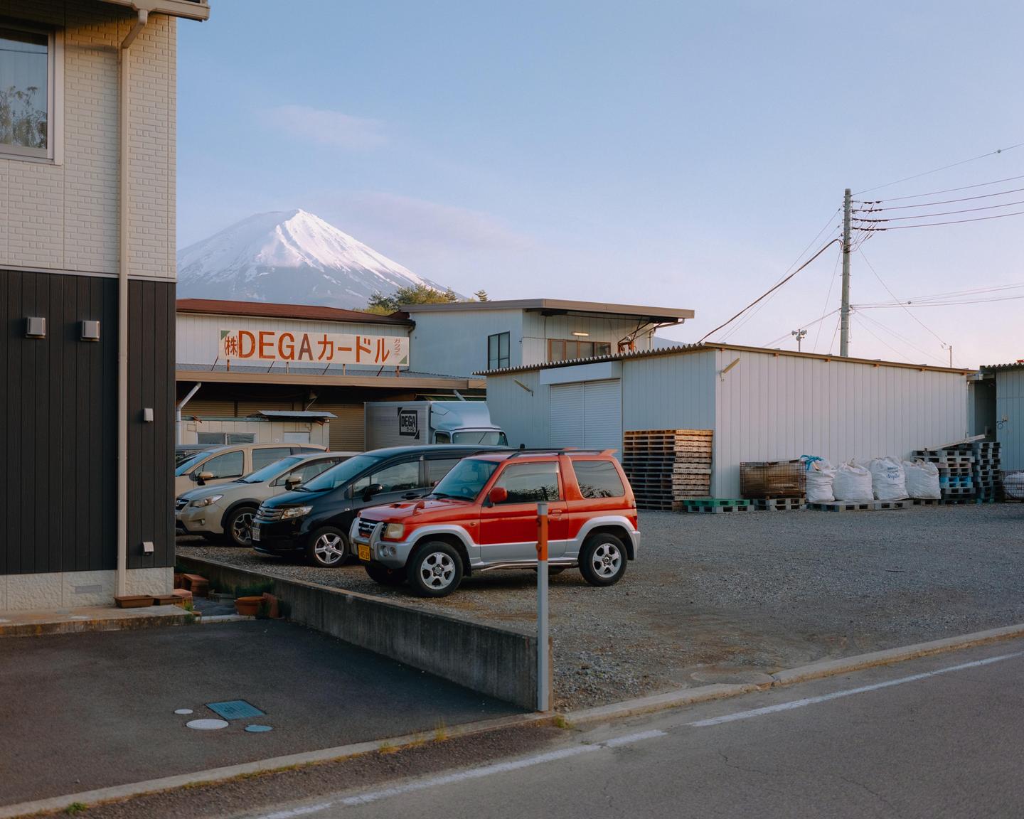 A snow-capped mountain rises above a parking lot with several cars and buildings.