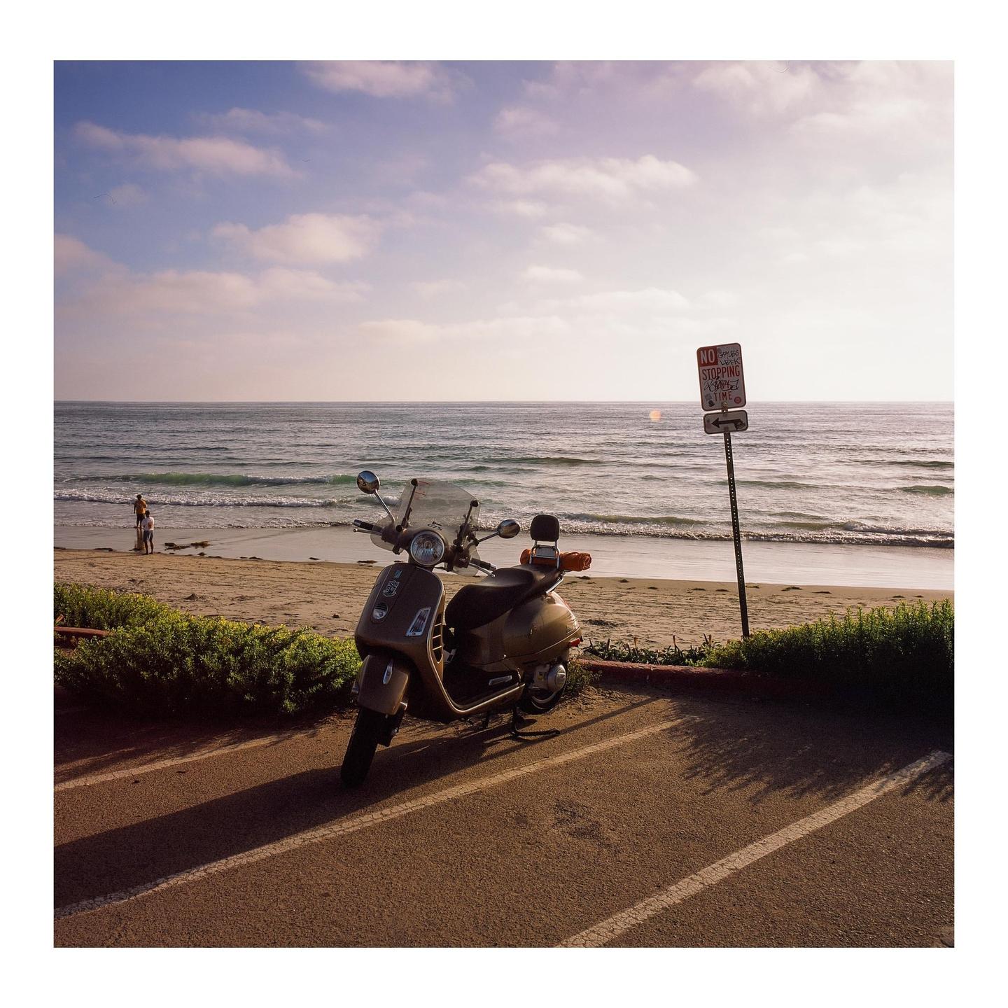 A scooter is parked on a paved area next to a sandy beach with the ocean in the background.