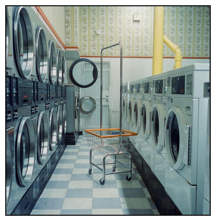 A laundry cart stands in the middle of a laundromat with rows of washing machines and dryers.