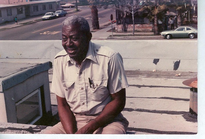 A man sits on a rooftop with a street and buildings in the background.