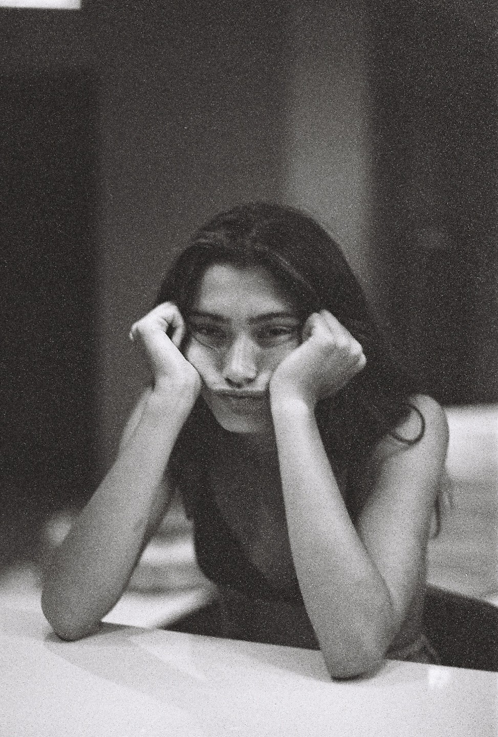A woman with long hair rests her chin on her hands and makes a pouting face.