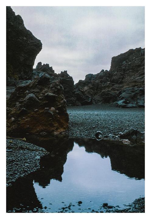 A pool of water reflects the sky and surrounding rock formations on a rocky beach.