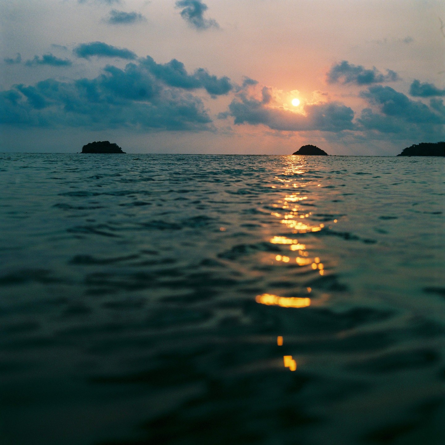 The sun sets over the ocean, casting a golden reflection on the water, with small islands visible in the distance.