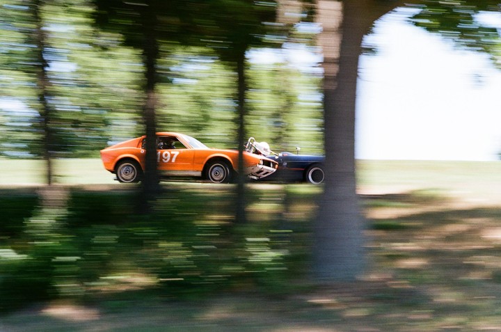 An orange and a dark blue race car speed past trees with blurred green foliage in the background.
