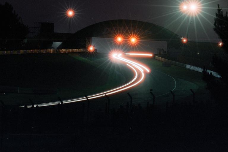Light trails from a car on a race track at night are visible under bright stadium lights.