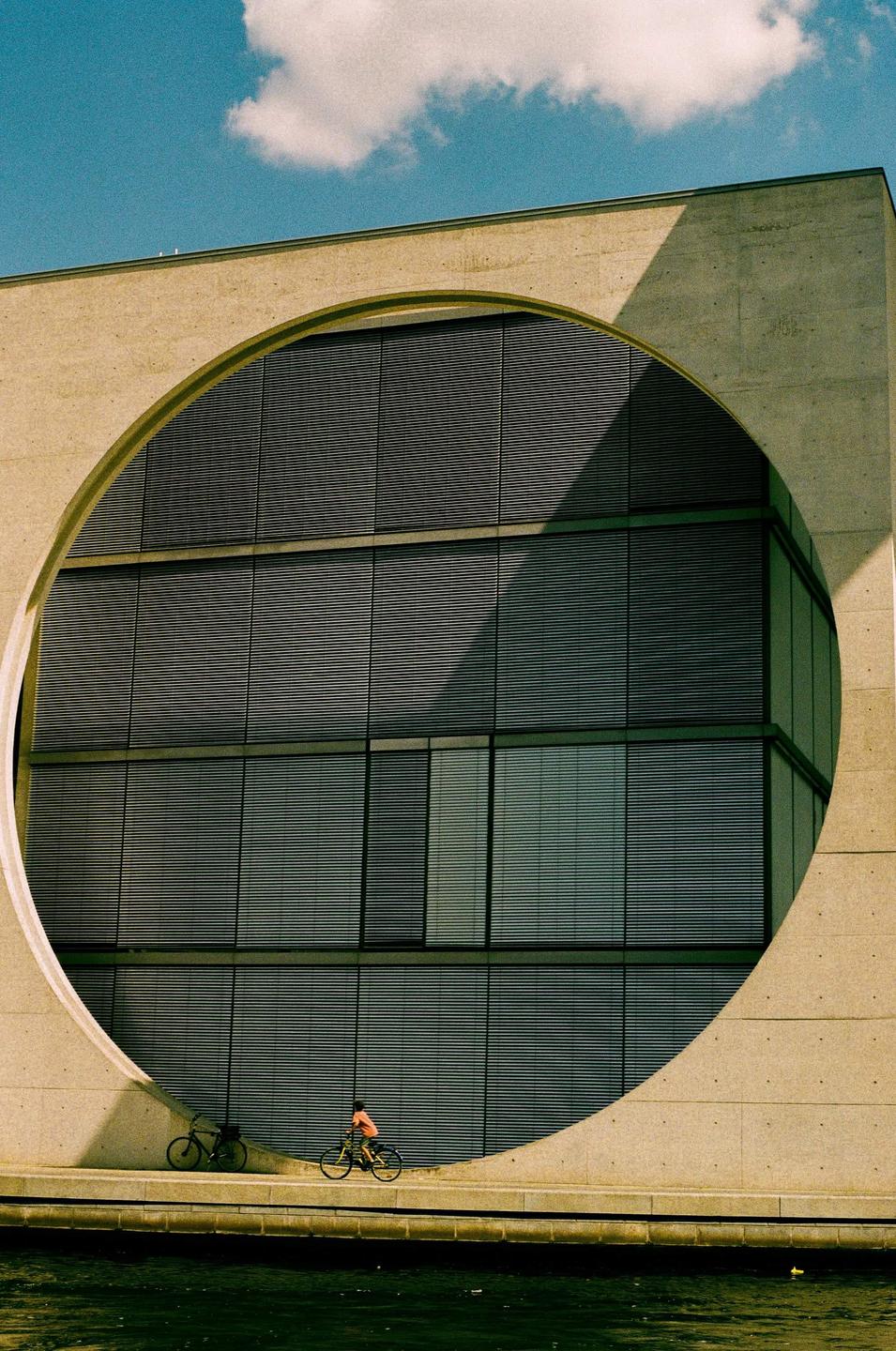 A cyclist rides past a large concrete building with a circular cut-out and many windows.