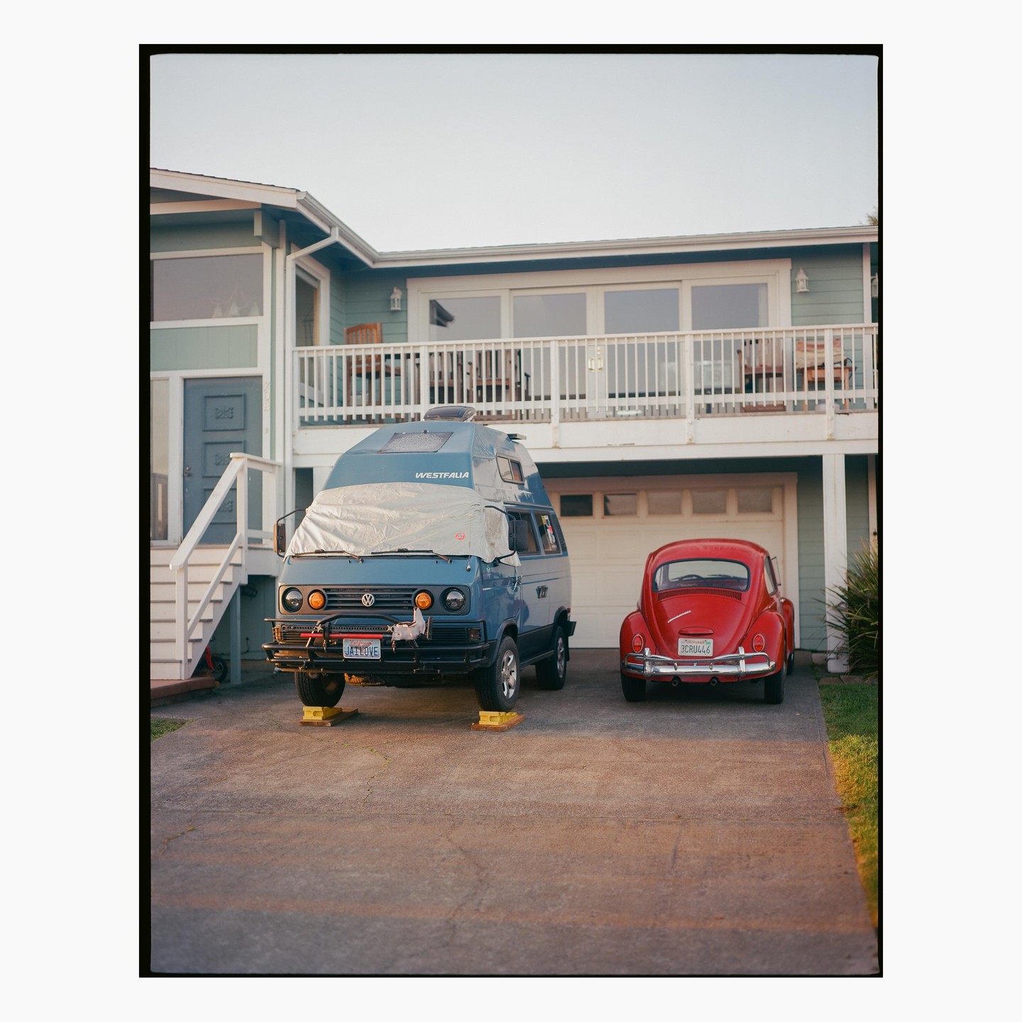 A blue camper van and a red beetle are parked in the driveway of a house with a balcony.