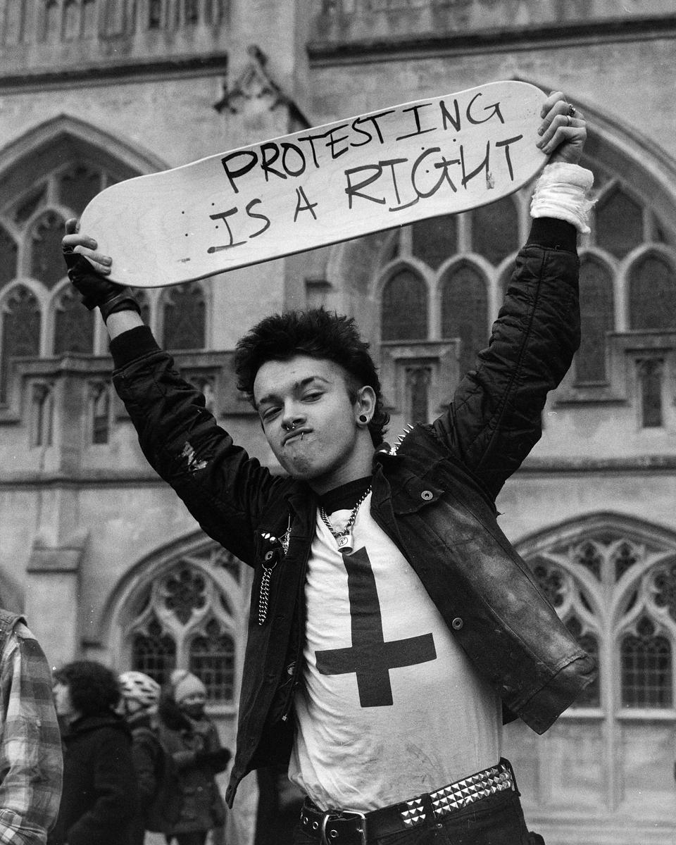 A young man with a skateboard over his head, with "PROTESTING IS A RIGHT" written on it, stands in front of a gothic building.
