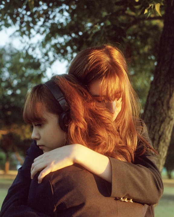 A woman with long red hair embraces a girl wearing headphones, with trees in the background.