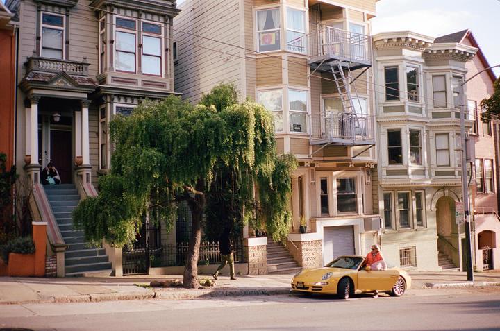 A yellow convertible car is parked on a street in front of several tall houses with a person sitting in the driver's seat.